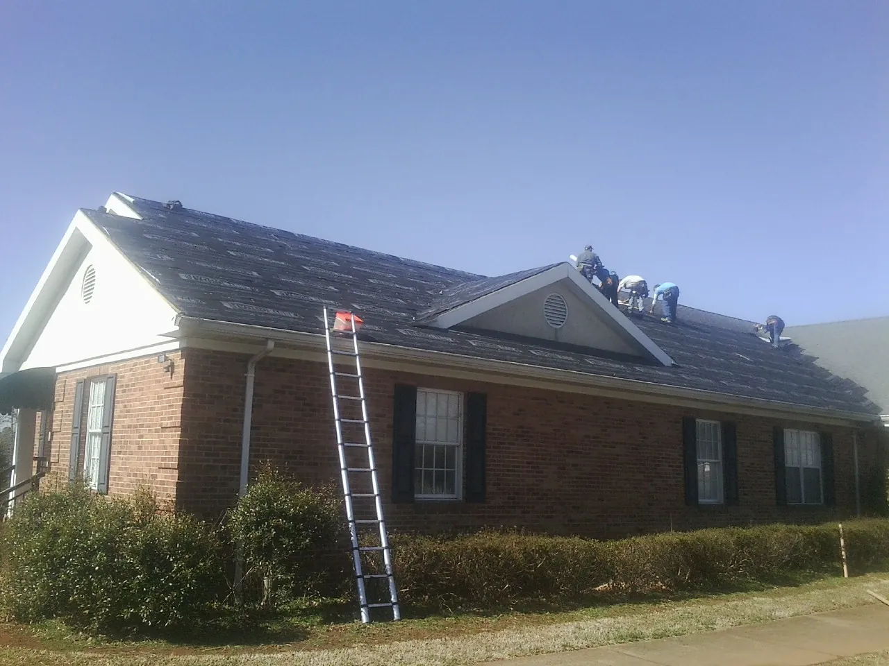 Crew tearing shingles off a brick ranch roof with a ladder set — Metal Roof Repair work by our crew in Willmar